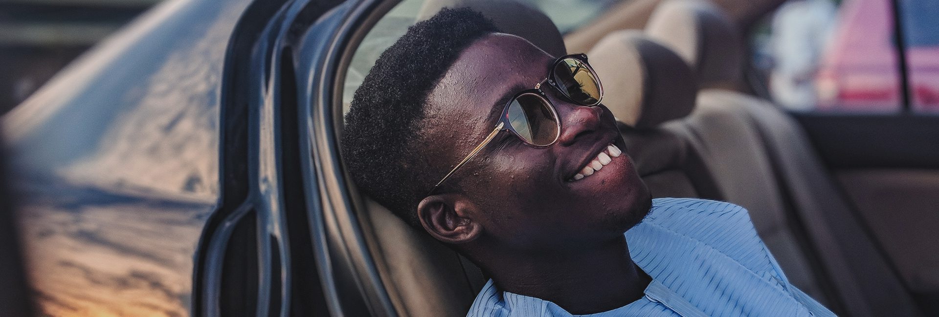 A man in sunglasses leans back in a car seat, looking up with a relaxed and happy demeanor.